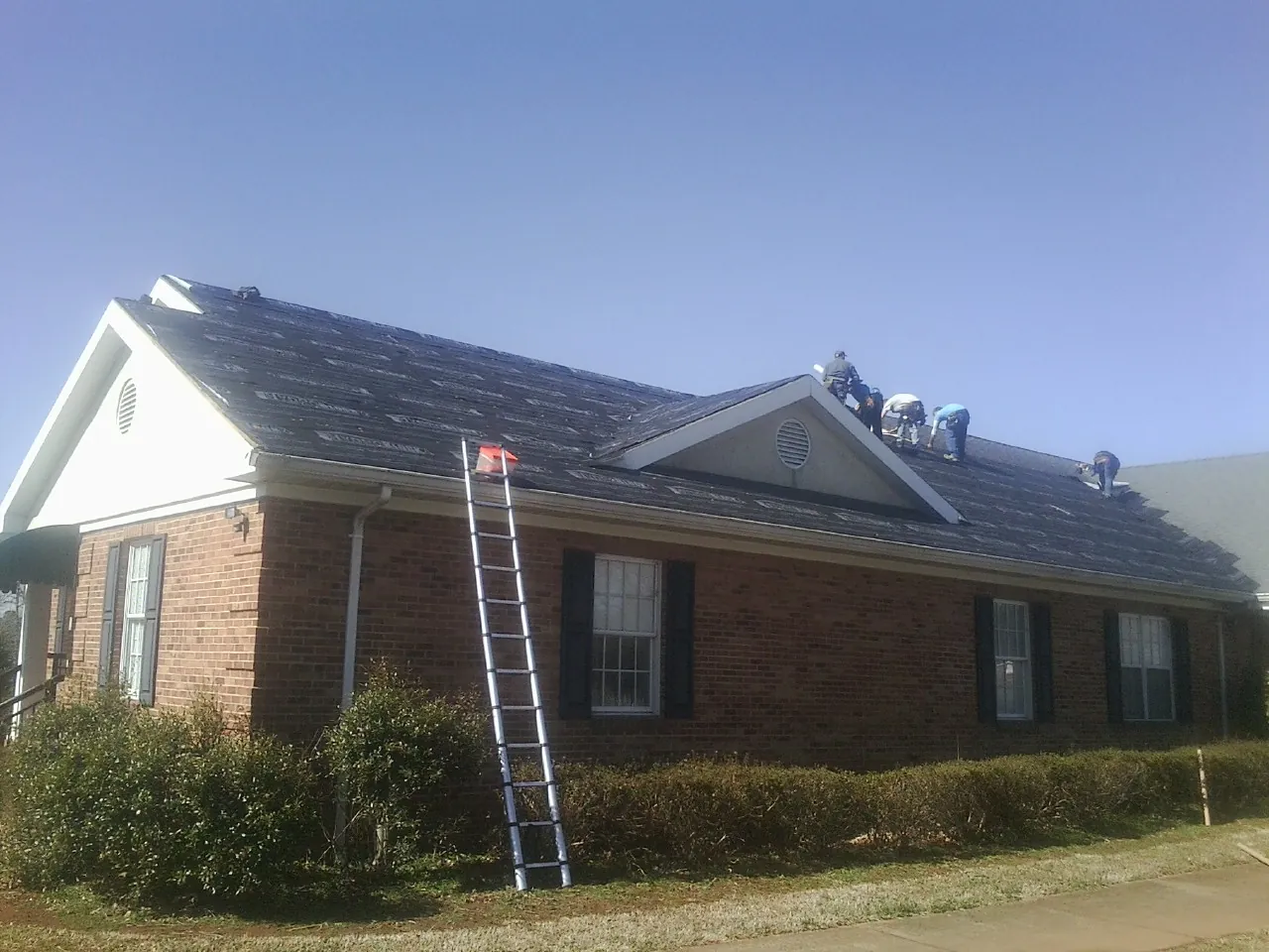 Crew tearing shingles off a brick ranch roof with a ladder set — Metal Roof Repair work by our crew in Indianola